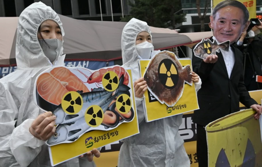 Aktivis lingkungan Korea Selatan yang mengenakan pakaian pelindung dan topeng (kanan) Perdana Menteri Jepang Yoshihide Suga memegang gambar ikan dengan tanda peringatan radioaktivitas saat protes terhadap keputusan Jepang melepaskan air limbah Fukushima, dekat kedutaan Jepang di Seoul Korsel pada Selasa 13 April, 2021.
