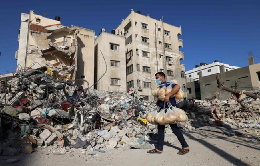Seorang pekerja toko roti Palestina yang membawa kantong roti melewati reruntuhan satu bangunan yang hancur selama pemboman Israel baru-baru ini, di Kota Gaza pada Kamis 27 Mei 2021.