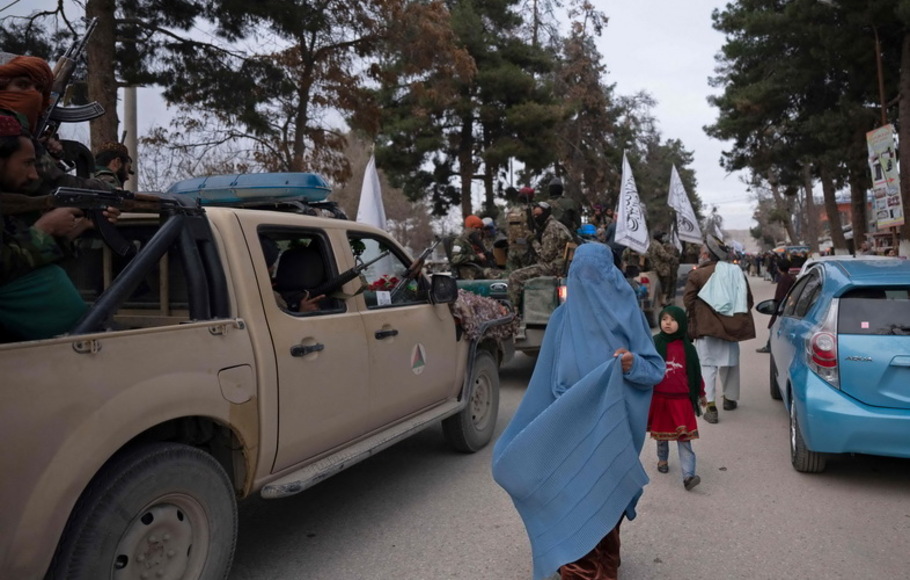 Seorang wanita Afghanistan berpakaian burqa berjalan melewati pejuang Taliban dengan kendaraan yang mengambil bagian dalam parade jalanan militer di Maymana, ibu kota provinsi Faryab, Minggu 16 Januari 2022. 