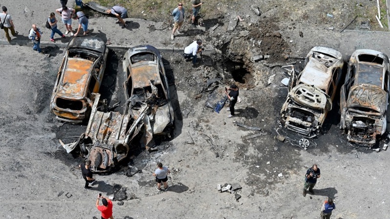 Sejumlah berkumpul di sekitar reruntuhan kendaraan dan kawah yang terbentuk setelah rudal Rusia menghantam halaman kompleks perumahan bertingkat di pinggiran timur Kharkiv, Ukraina pada 26 Juni 2022 di tengah serangan Rusia ke Ukraina. (FOTO: SERGEY BOBOK / AFP)