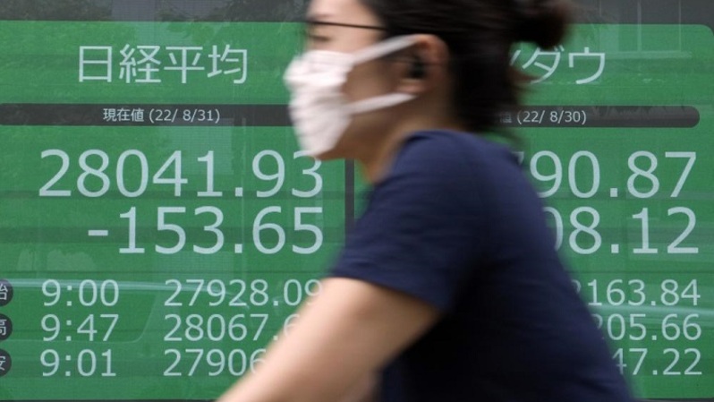 Seseorang yang memakai masker pelindung mengendarai sepeda di depan papan saham elektronik yang menunjukkan indeks Nikkei 225 dan Dow New York di sebuah perusahaan sekuritas pada 31 Agustus 2022 di Tokyo, Jepang. (FOTO: AP/Eugene Hoshiko)