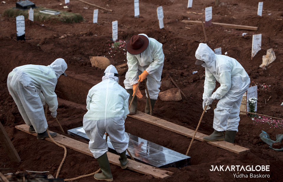 Funeral service workers wearing protective gear lower the plastic-wrapped coffin into the grave. (JG Photo/Yudha Baskoro)