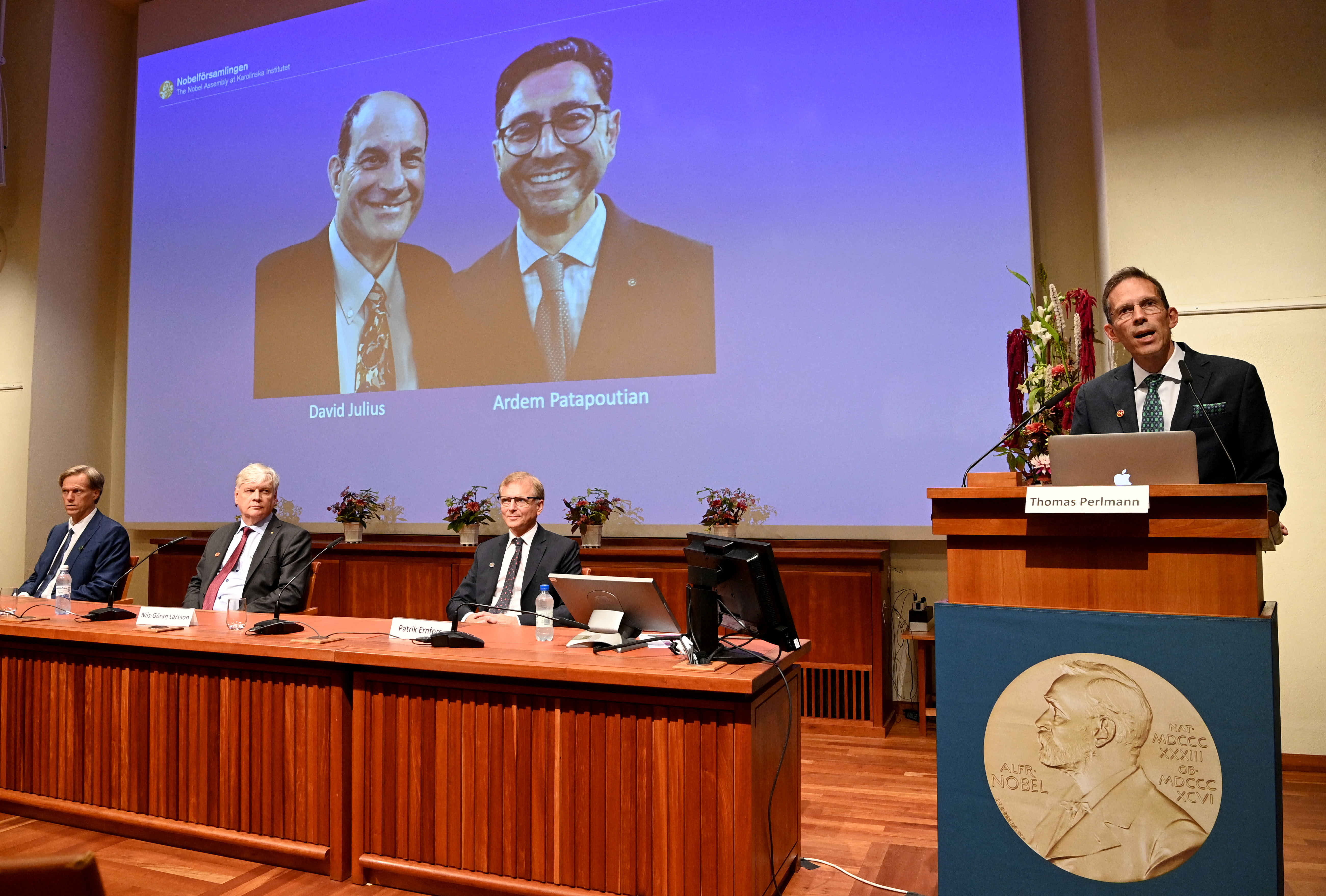 Sekretaris Komite Nobel, Thomas Perlmann (kanan) berdiri di samping layar yang memperlihatkan  pemenang Hadiah Nobel bidang Fisiologi atau Kedokteran 2021, David Julius (kiri) dan Ardem Patapoutian, selama konferensi pers di Institut Karolinska di Stockholm, Swedia, pada 4 Oktober 2021.  ( Foto: JONATHAN NACKSTRAND / AFP )